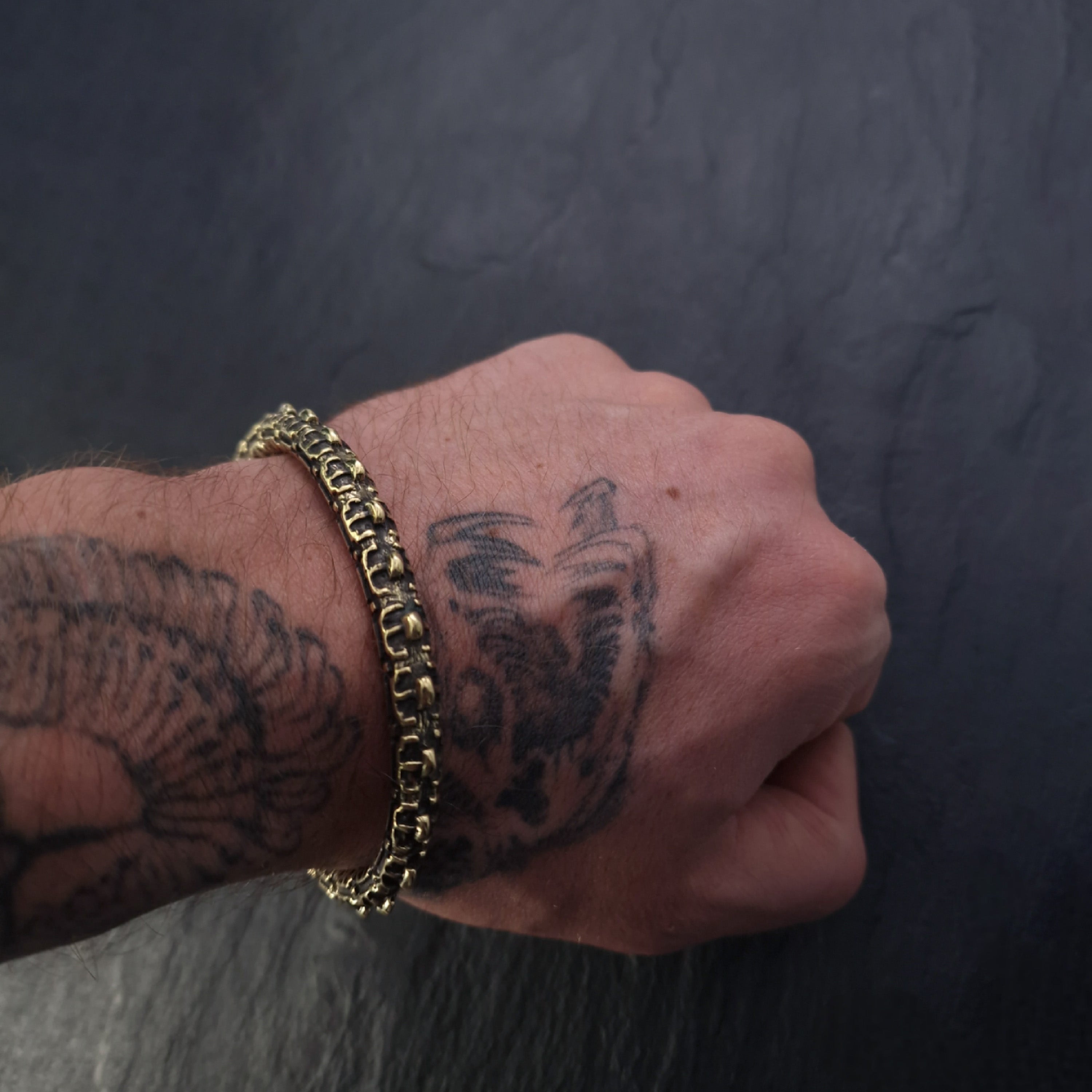 A person's wrist, adorned with a visible tattoo of an eagle, is wearing the SHIRO Men's Gold Cuff Bracelet by Kaya Movement. The background features a dark, textured surface.